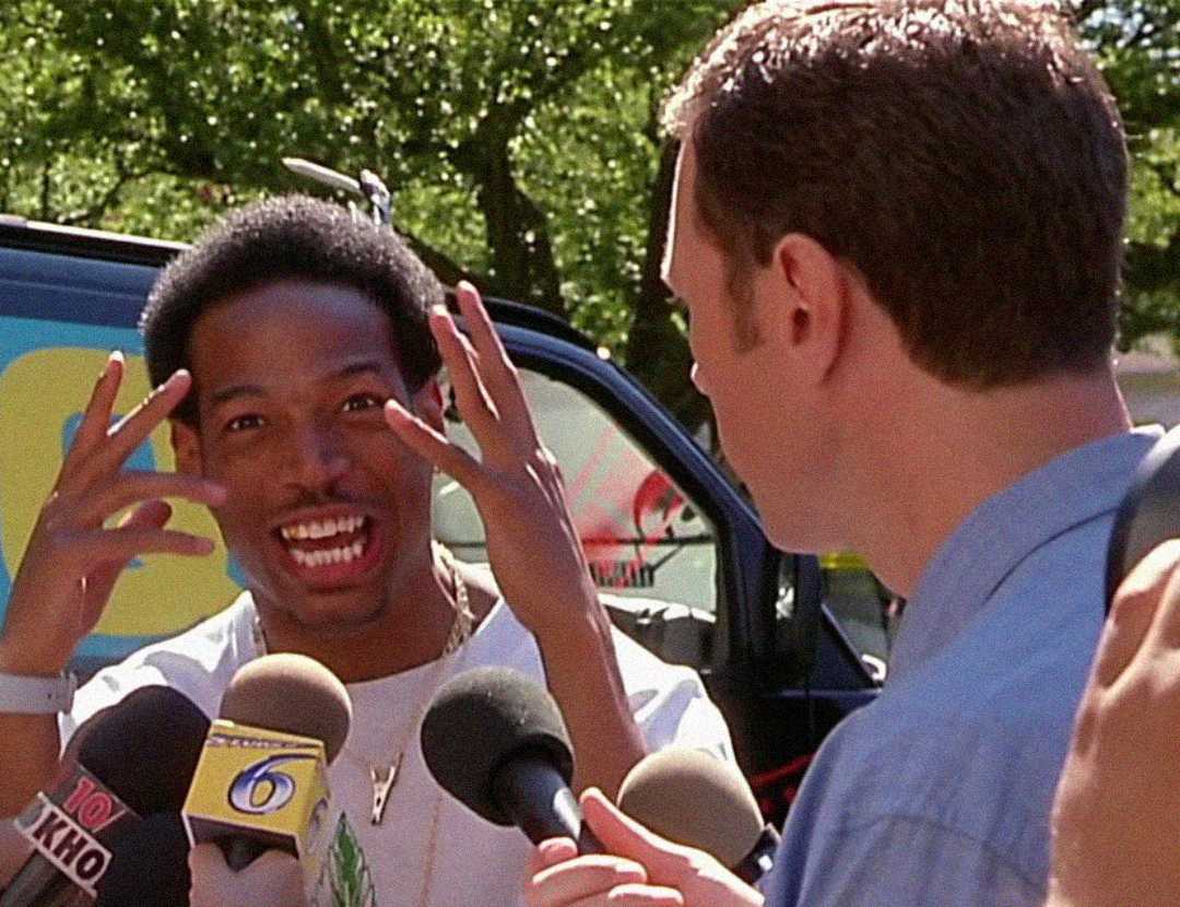 A man animatedly gestures with his hands while speaking to reporters holding microphones. Another man, possibly a journalist, faces him. A news van and trees are visible in the background.