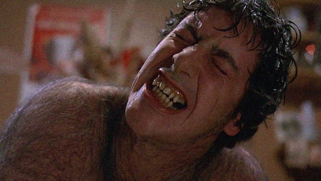 A man with messy hair, yellowed teeth, and thick body hair grimaces in pain, his face contorted and eyes tightly shut, suggesting he is undergoing a painful transformation. The background is blurry and indoors.