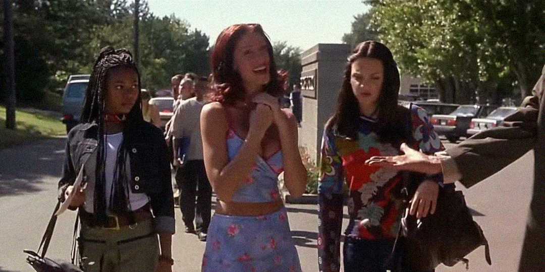 Three young women walk together outside on a sunny day, talking and smiling. Trees, cars, and other people are visible in the background as they stroll along a sidewalk.