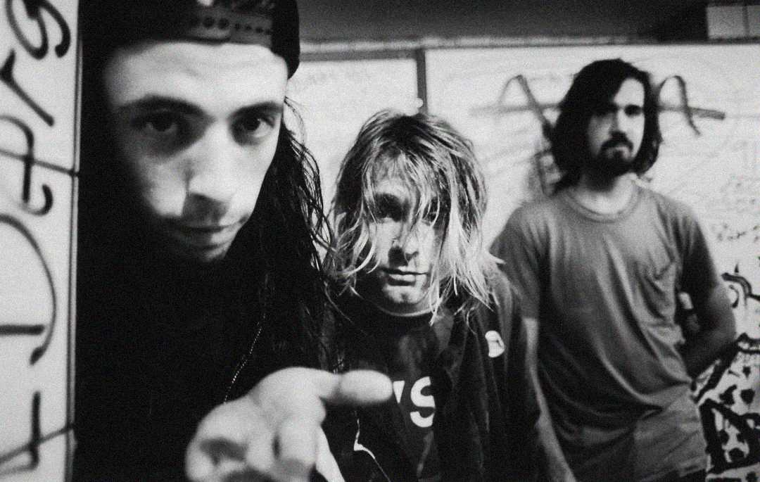 Three men stand in a grungy, graffiti-covered room. The man in front wears a backwards cap and reaches toward the camera. The man in the middle has messy blonde hair, and the third has dark hair and a beard. The image is black and white.