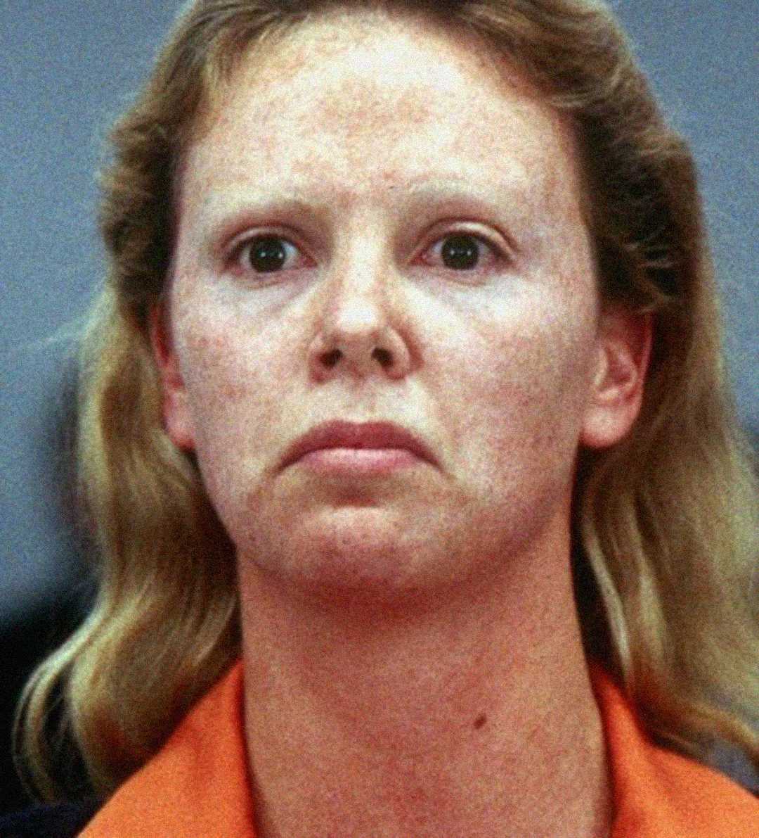 A close-up of a person with light hair and pale eyebrows, wearing an orange collared shirt, looking forward with a neutral expression.