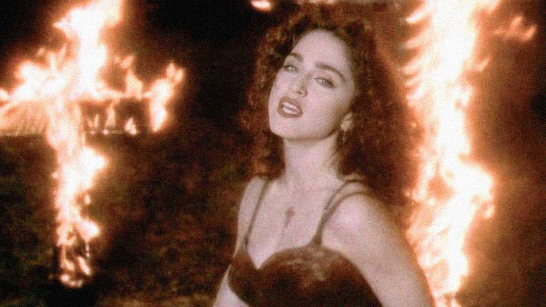 A woman with curly dark hair and red lipstick stands in front of large burning crosses at night, wearing a black dress and a necklace. The scene is dramatic and intense with flames illuminating the background.