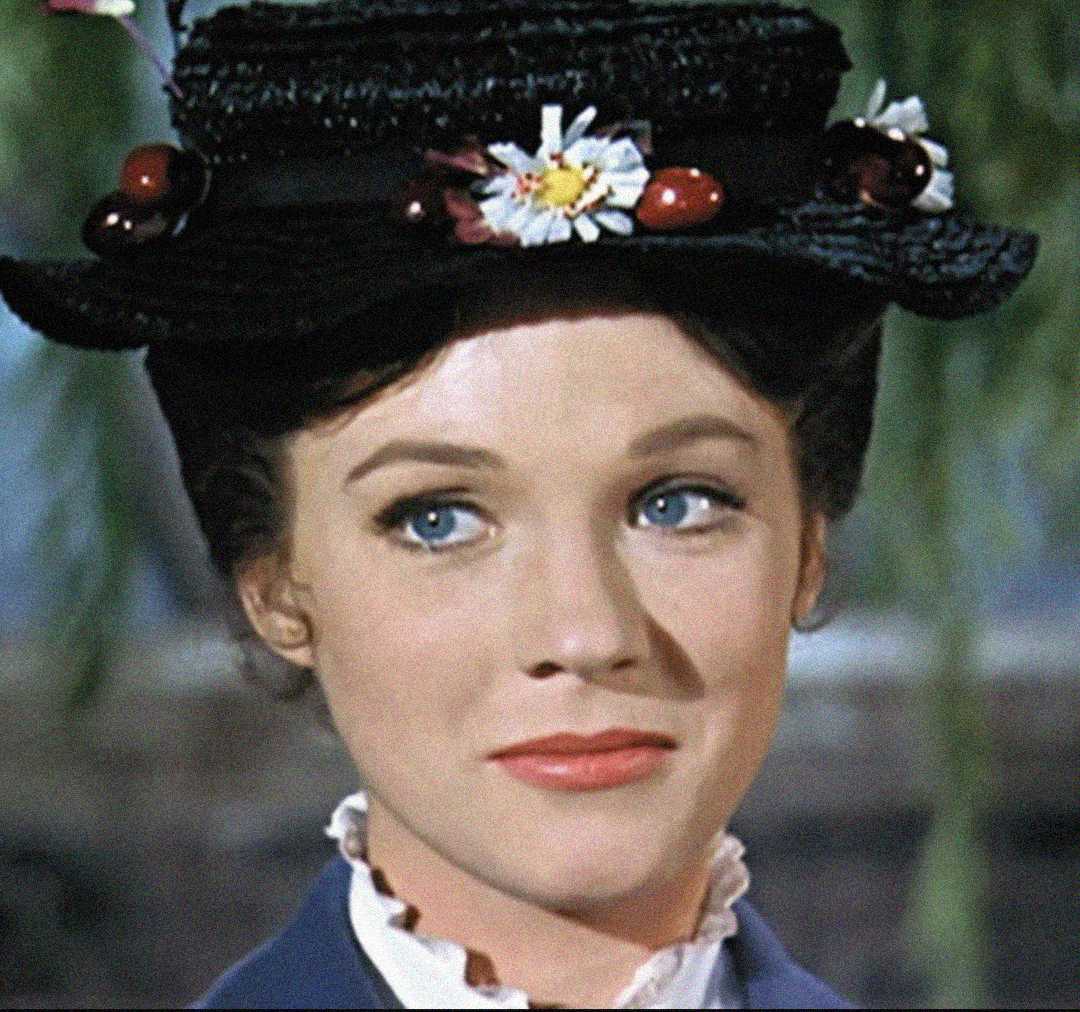 A woman with blue eyes and light makeup wears a dark hat decorated with flowers and cherries, and a ruffled white collar, looking slightly to the side with a gentle expression.
