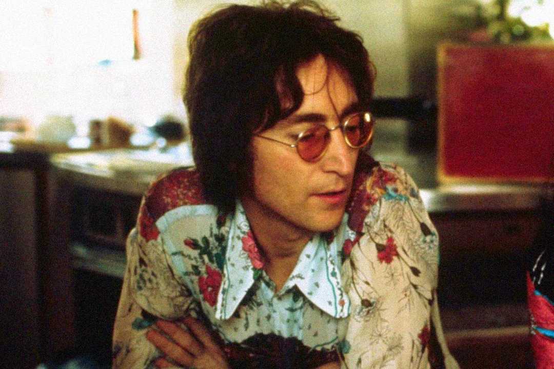 A man with wavy dark hair and round glasses sits with arms crossed, wearing a floral patterned shirt. He appears thoughtful and is looking down in a softly lit indoor setting.