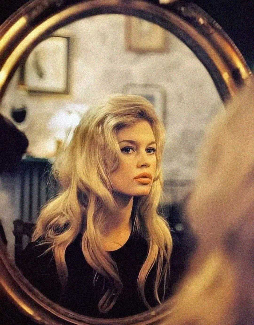 A woman with long blonde hair is seen through the reflection of a round mirror, gazing thoughtfully. The background is softly blurred with framed pictures and light decor visible.