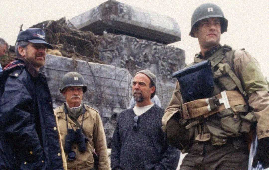 Four men stand together outdoors; two wear WWII-era military uniforms and helmets, while the other two wear modern clothing and hats. A large, weathered structure is in the background. The mood is serious and focused.