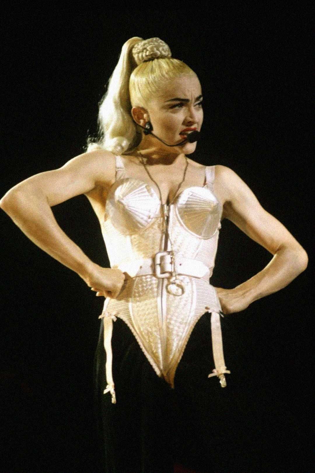 A performer with platinum blonde hair in a high ponytail wears a cream-colored, cone-shaped corset top and black pants on stage, striking a confident pose under bright lighting.
