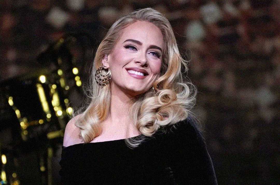 A woman with long blonde hair styled in waves, wearing a black off-the-shoulder dress and large gold earrings, smiles while looking slightly to the side. The background is blurred with warm lighting.