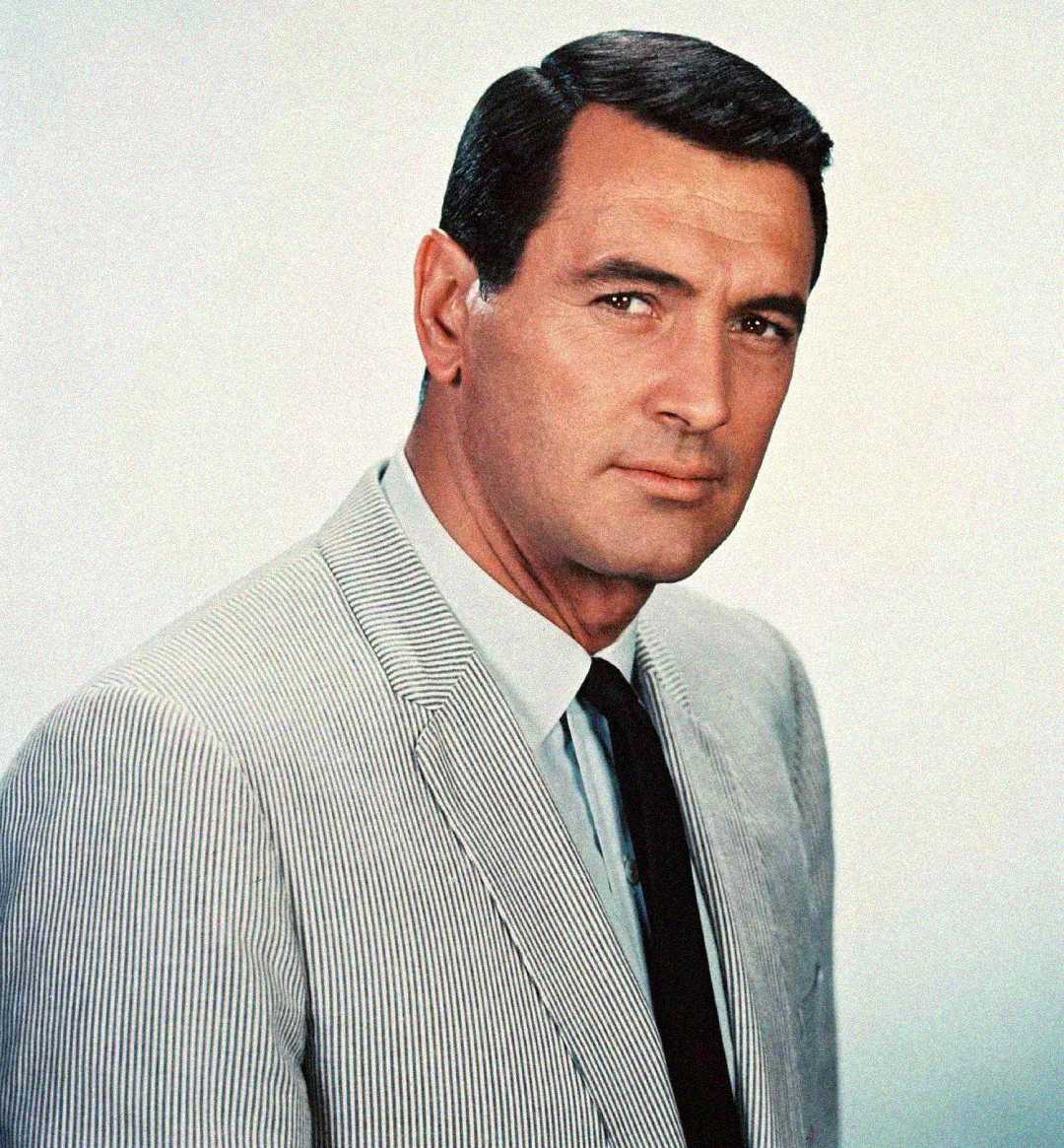 A man with dark hair, wearing a light-colored pinstripe suit, white shirt, and dark tie, poses against a plain light background, looking confidently at the camera.