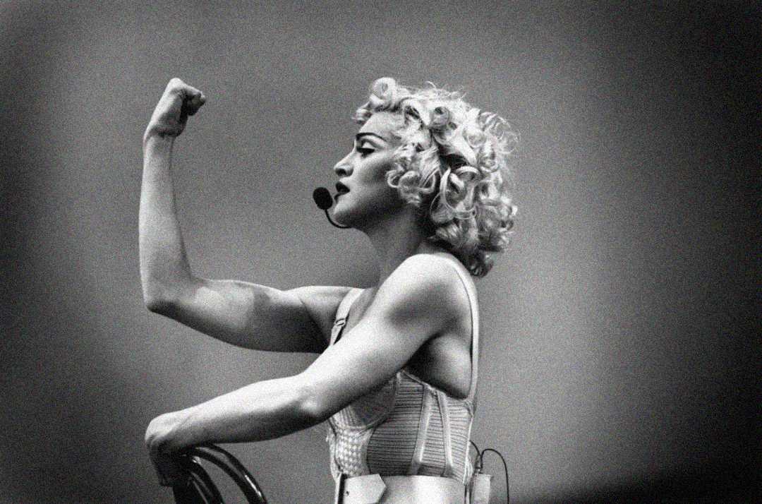 Black and white photo of a person with curly blonde hair, wearing a headset microphone and a cone-shaped corset, flexing their arm in a strong pose while looking to the side.