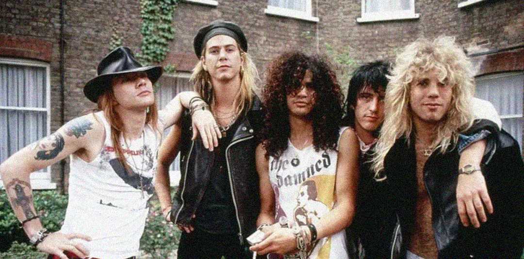 Five men with long hair and rock-style clothing pose together outdoors in front of a brick building with windows. They wear leather vests, hats, and band shirts, with some arms draped over each other's shoulders.