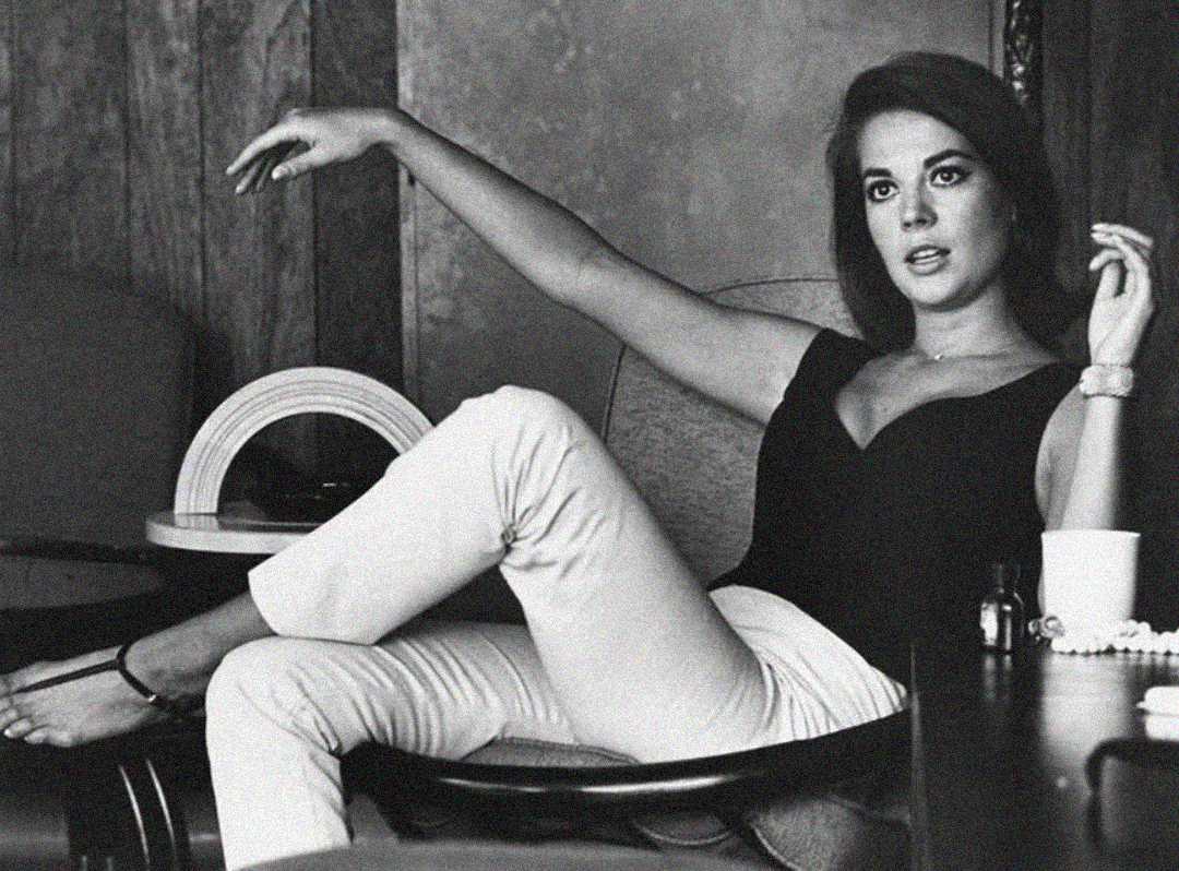A woman in a sleeveless black top and light pants lounges in a chair with one arm raised and the other resting on the chair's back, looking off-camera. A cup, bottle, and decorative object sit on the table nearby.