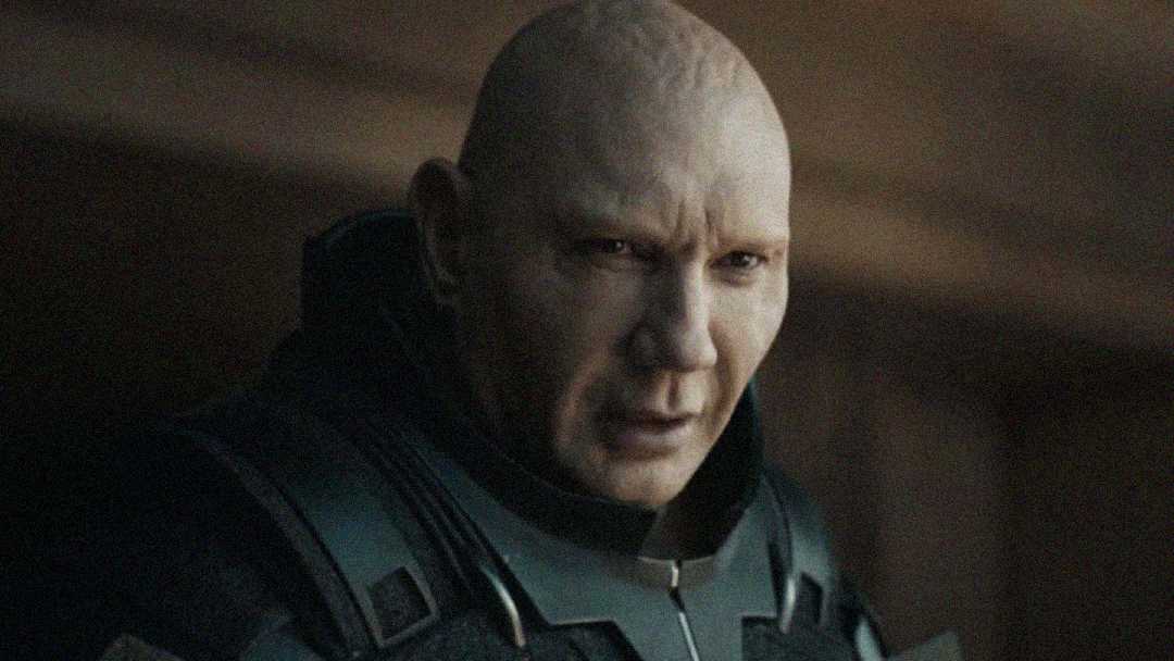 A bald man in dark, futuristic armor looks intently ahead with a serious expression. The lighting is dim and the background is blurred, drawing focus to the man's face and detailed armor.