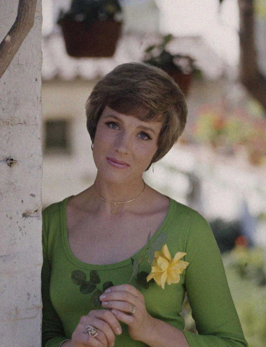 A woman with short brown hair, wearing a green top and a necklace, stands outdoors holding a yellow rose and looking gently at the camera. There are hanging potted plants and blurred flowers in the background.