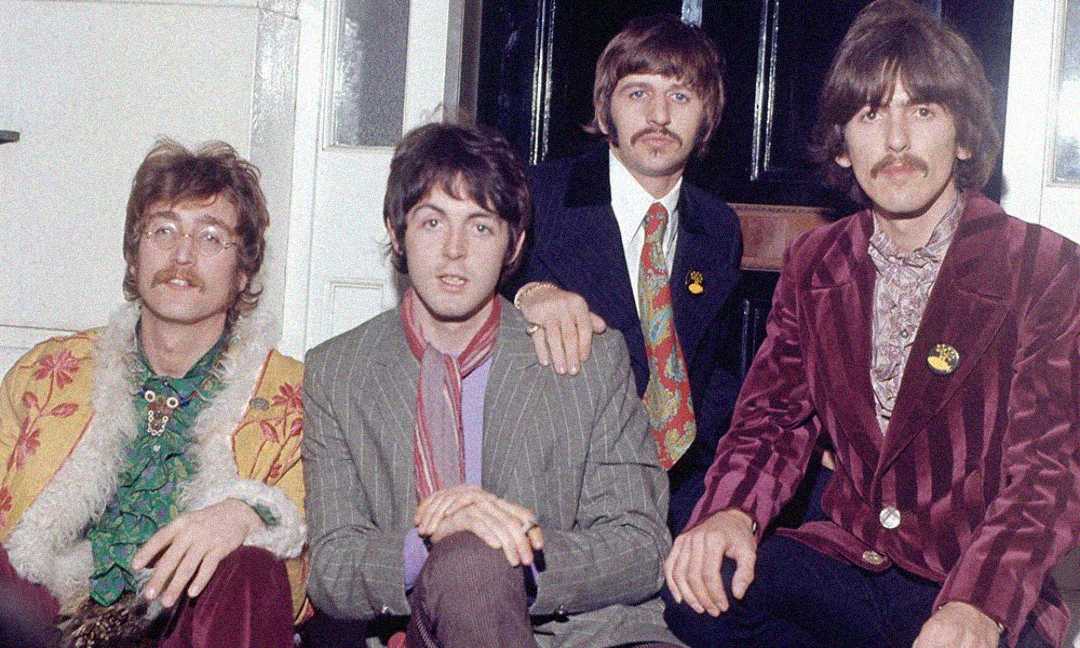 Four men with 1960s-style hair and clothing sit together on steps. They wear colorful, patterned jackets and shirts. Three have mustaches, and all look toward the camera. The setting appears to be outside a building.