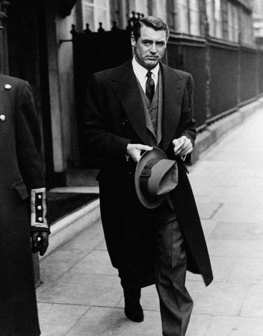 A man in a long overcoat and suit walks confidently down a city sidewalk, holding a fedora hat in one hand. The image is black and white, and the street and railings are visible in the background.