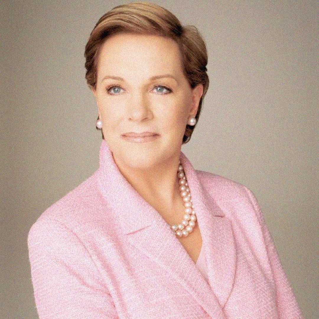 A woman with short light brown hair wears a light pink blazer, double-stranded pearl necklace, and pearl earrings, looking slightly to the side against a plain light background.