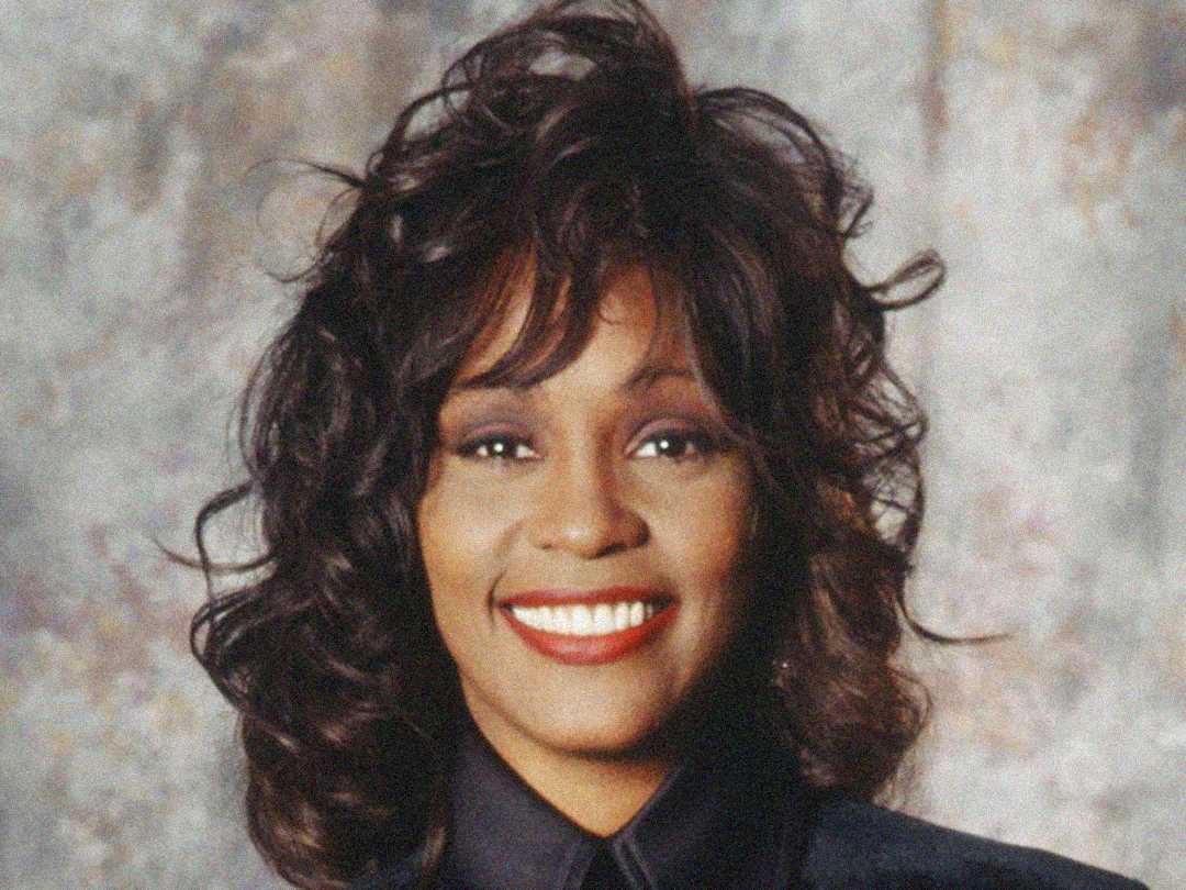 A woman with medium-length, wavy dark hair smiles brightly at the camera. She is wearing a dark top and has a light background behind her.