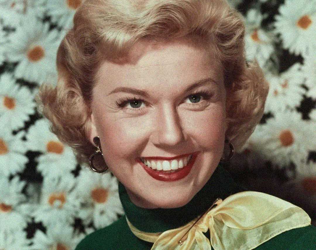A woman with short blonde, curly hair smiles brightly. She wears hoop earrings, a green top with a large yellow bow on the shoulder, and stands in front of a background filled with white daisies.