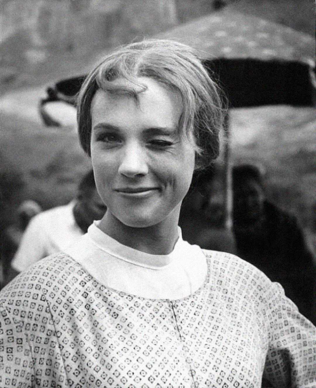 A young woman with short hair winks at the camera, smiling slightly. She wears a patterned top and stands outdoors with an umbrella and blurred figures in the background. The image is in black and white.