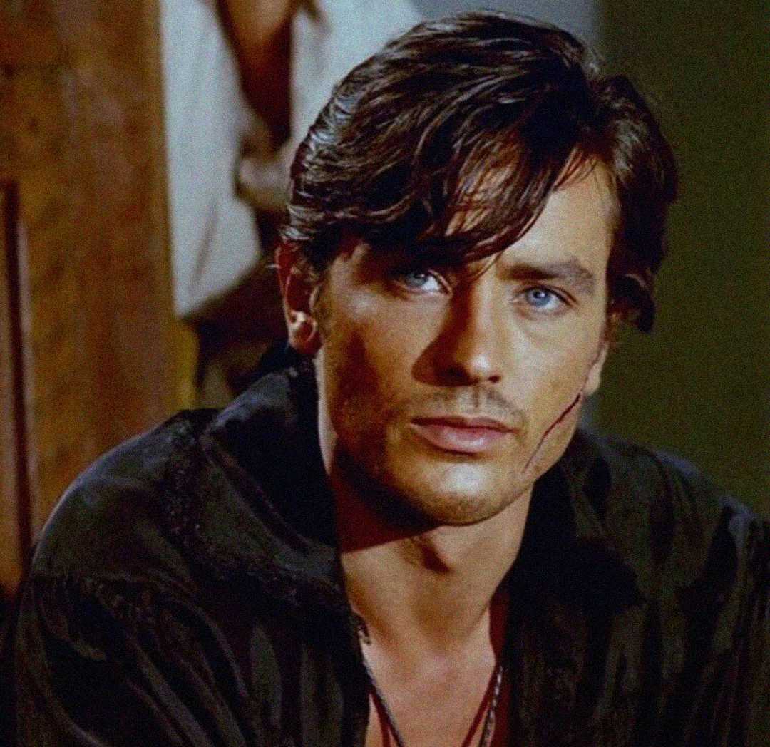A man with tousled dark hair and blue eyes wears a dark shirt and has a faint scar on his cheek, looking seriously toward the camera in warm lighting.
