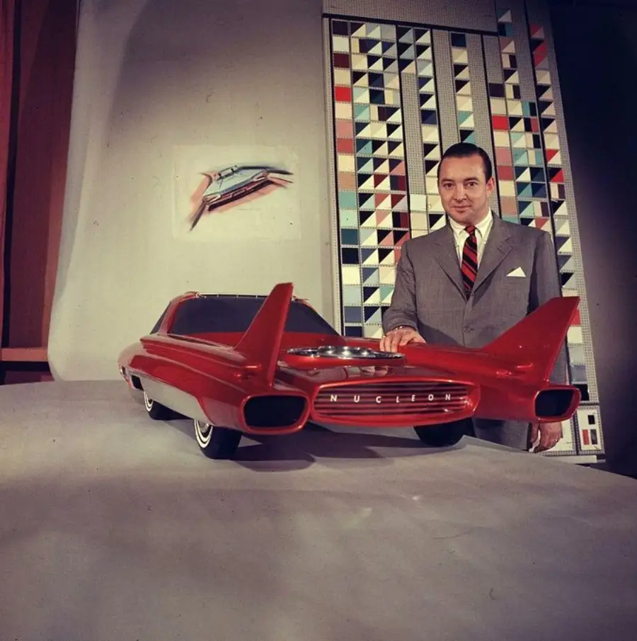 A man in a suit stands beside a futuristic red car model with large tailfins. Behind him is a geometric patterned wall and a drawing of a car on the wall.
