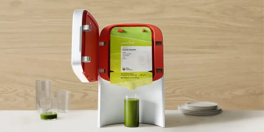 A countertop juicer with an open lid holds a green juice pouch labeled "Sweet Greens." Below, a glass is being filled with green juice. There are additional empty glasses and stacked white plates on the wooden surface.