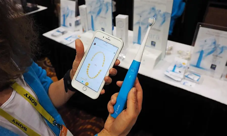 A person holds a blue electric toothbrush in one hand and a smartphone showing a dental app with a diagram of teeth in the other, at an exhibition booth with dental product displays in the background.