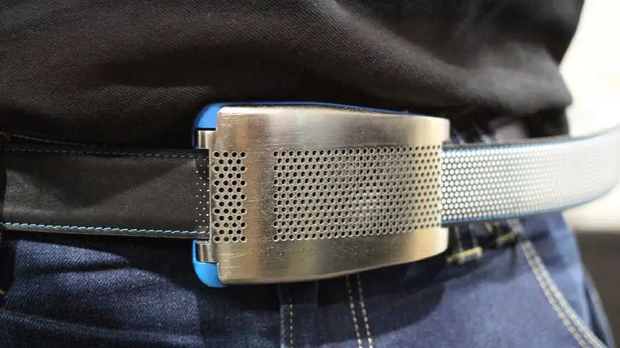 A close-up of a person wearing a metallic smart belt with a perforated, rectangular buckle, paired with dark blue jeans and a black shirt.