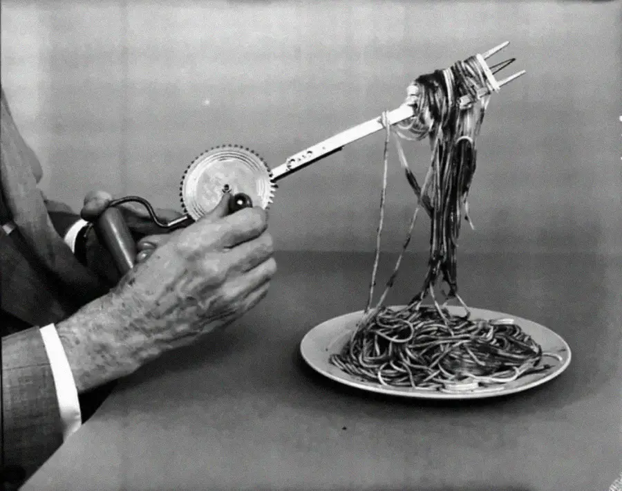 A person uses a hand-cranked mechanical fork to twirl spaghetti from a plate, demonstrating an unusual and humorous way to eat pasta.