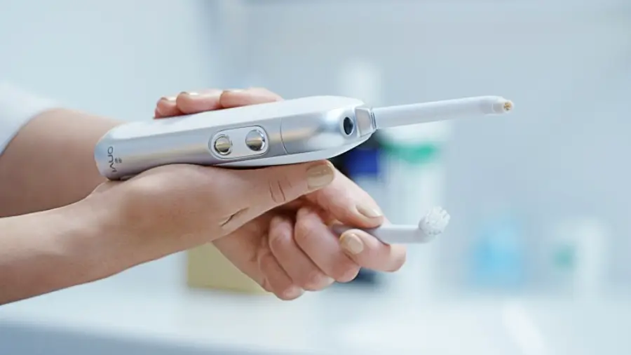 A person holds an electric toothbrush, attaching or detaching the brush head in a bathroom setting with blurred toiletry items in the background.