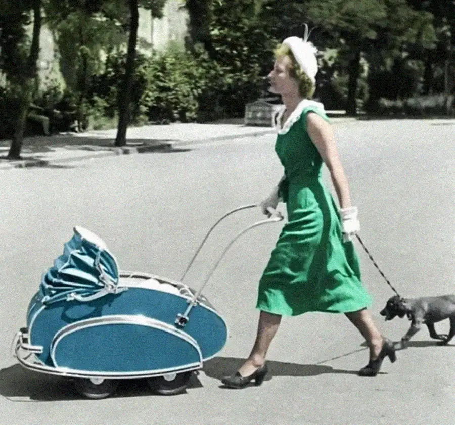 A woman in a green dress, white gloves, and a hat walks outdoors, pushing a vintage blue baby carriage and holding a small dog on a leash. Trees and pavement are visible in the background.