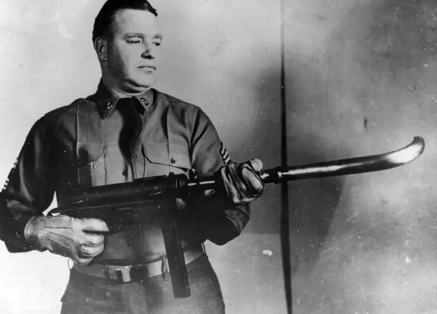A man in a military uniform stands holding a submachine gun with an unusually long, curved attachment at the end of the barrel, posing for the camera in a black-and-white photo.