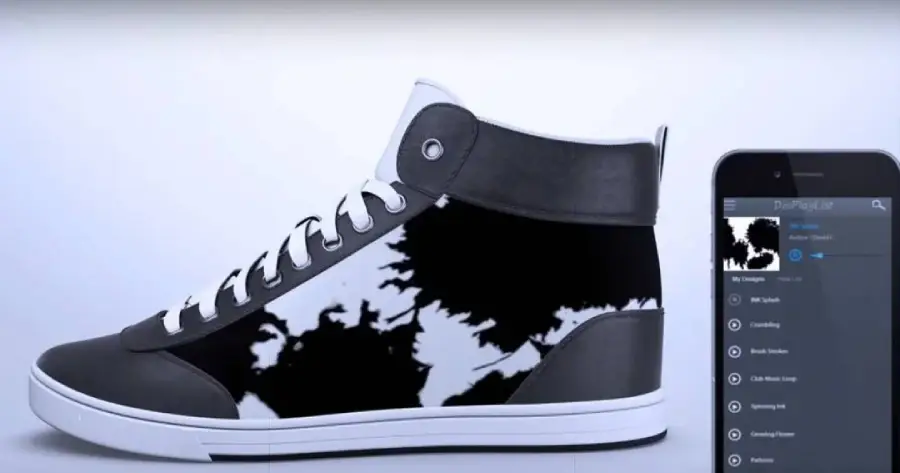 A black and white high-top sneaker with a digital display on its side shows an abstract pattern. Next to the shoe, a smartphone screen displays an app interface for customizing the shoe’s design.