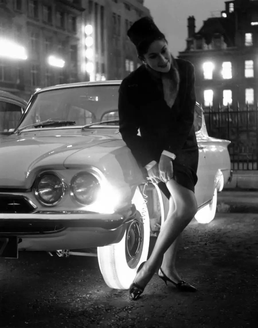 A woman in a dark dress leans casually against the front of a vintage car with glowing whitewall tires, on a city street at night, surrounded by illuminated buildings.
