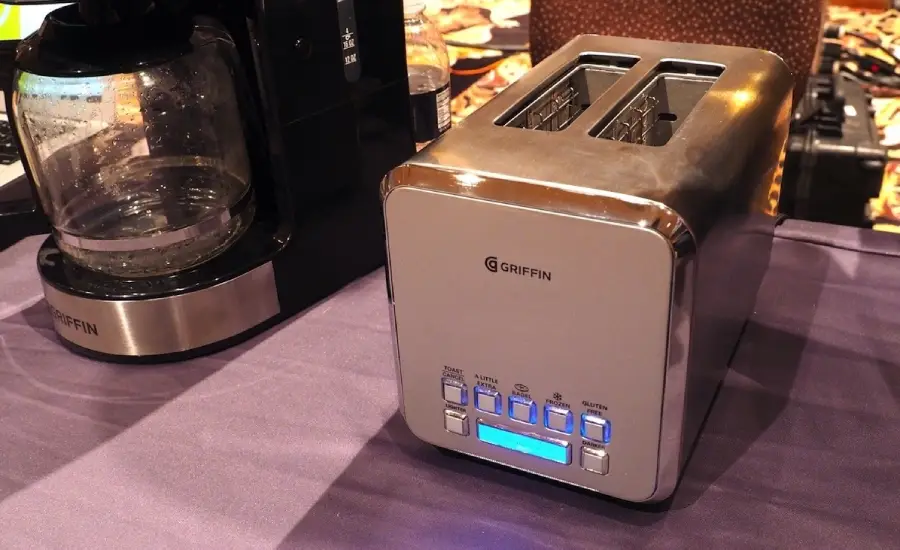 A silver Griffin toaster with digital controls and a blue display sits on a purple tablecloth next to a coffee maker with a glass carafe.