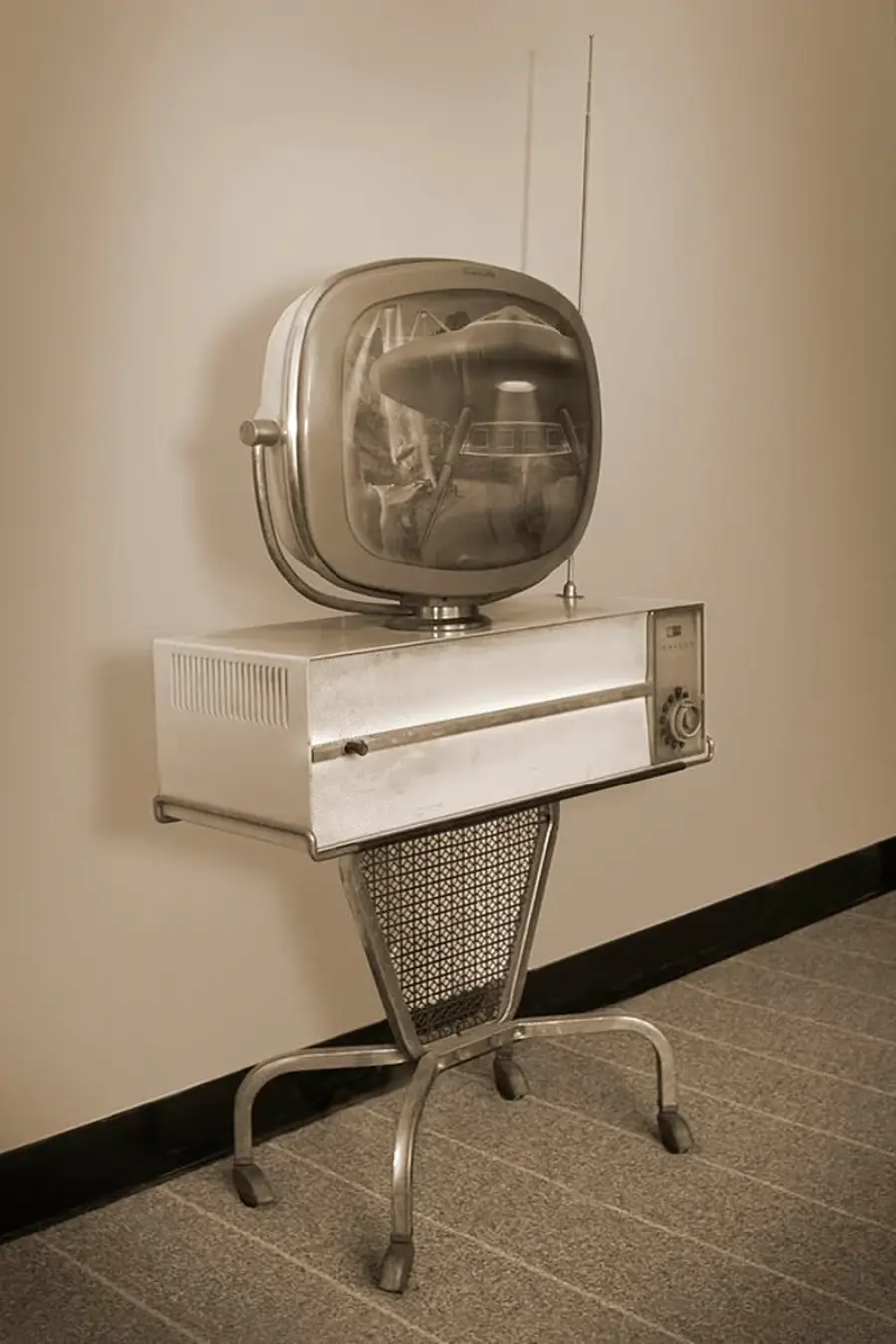 A vintage television with a rounded screen, antenna, and metal stand is positioned against a plain wall on a carpeted floor. The image is in sepia tones, giving it an old-fashioned look.