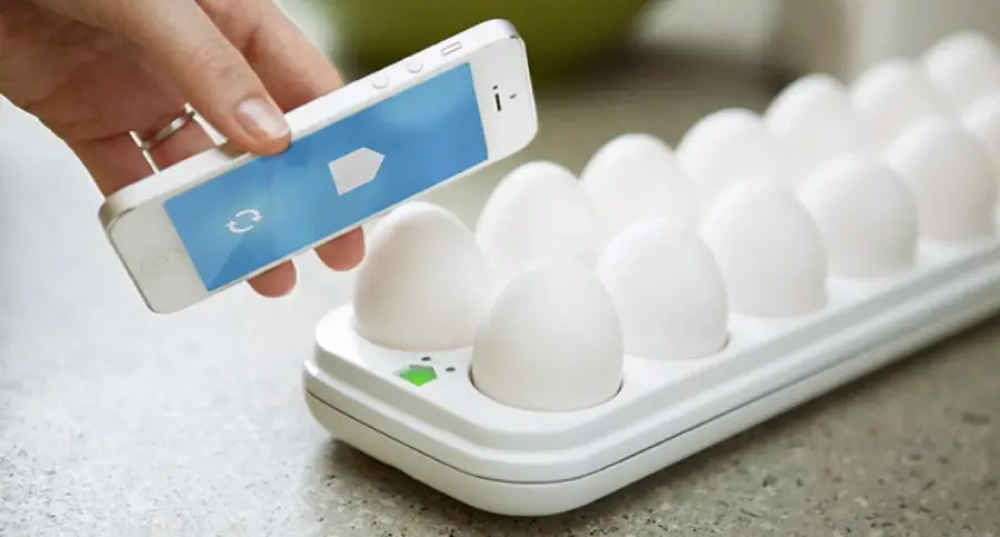 A hand holds a smartphone with a blue screen over a white egg tray containing several white eggs, suggesting the use of a smart device to monitor the eggs.