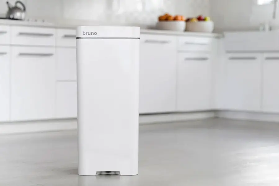A sleek, white kitchen trash can labeled "bruno" stands on a clean, light-colored floor with modern white cabinets and a bowl of fruit in the background.