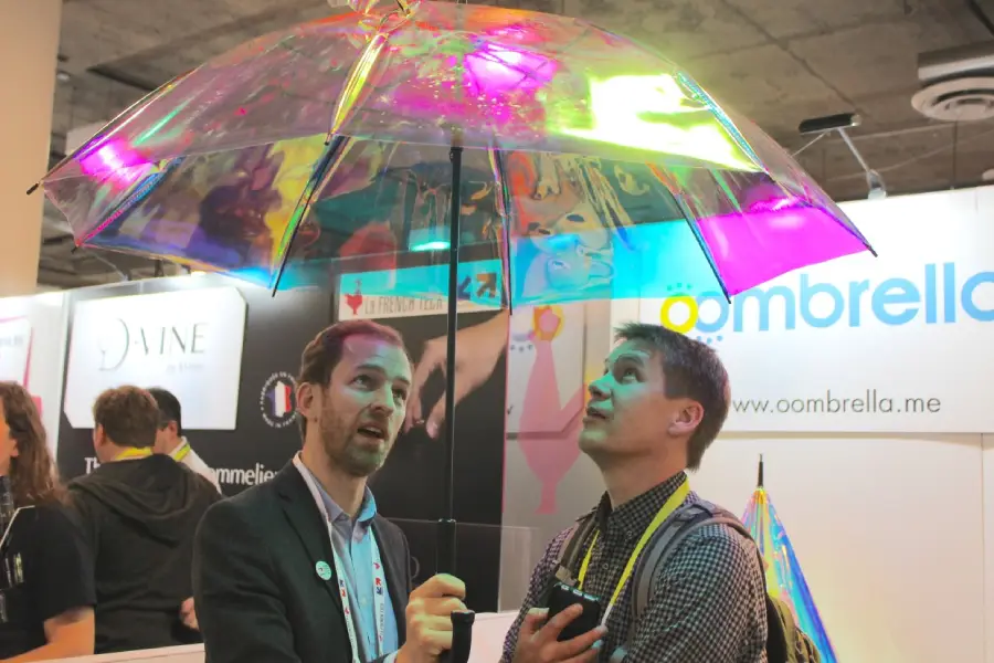 Two men stand indoors under a transparent, colorful umbrella, looking up at it. They appear curious and engaged. Behind them are exhibition booths and a sign with the word "Oombrella.