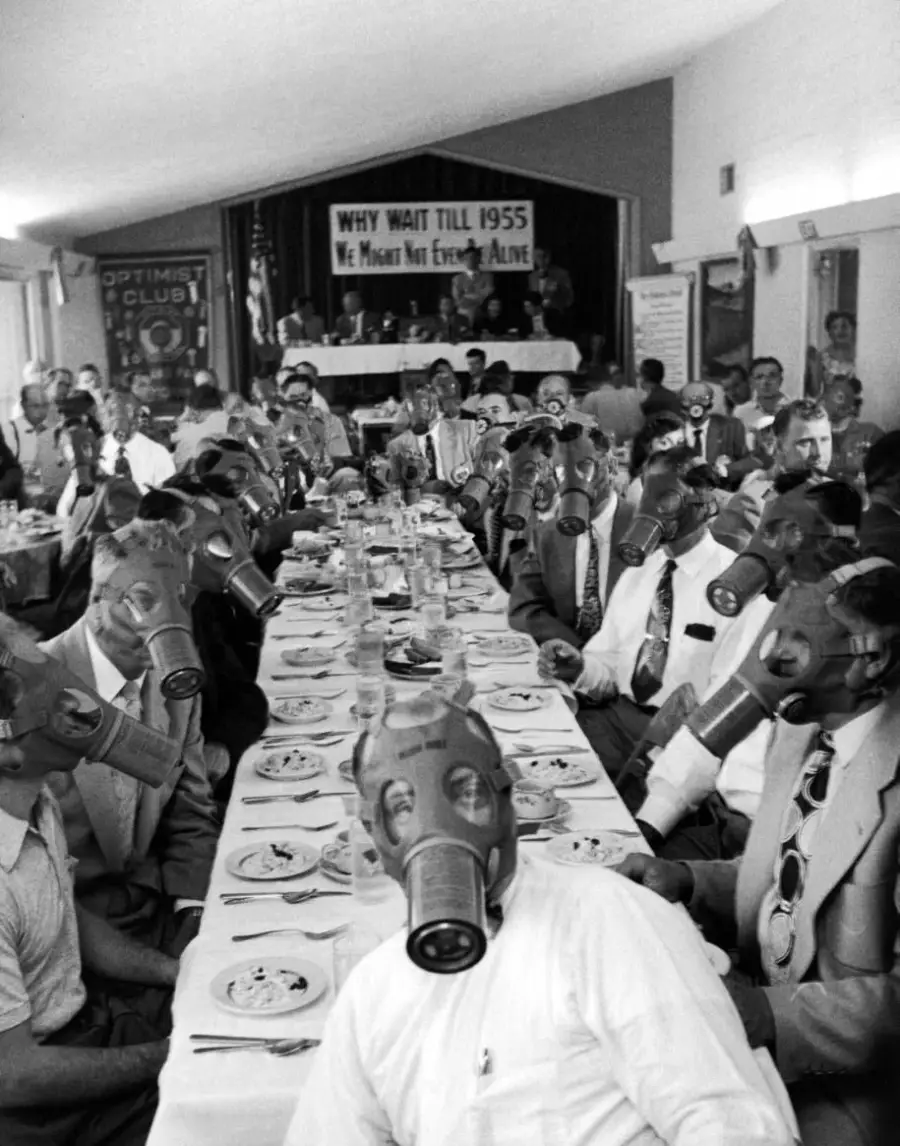 A large group of men wearing suits and gas masks sit at long dining tables, eating a meal. A banner on the wall reads, “Why wait till 1955, we might not even be alive.” Several people sit at a head table behind them.