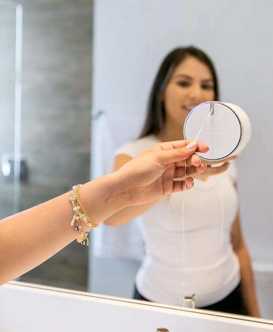 A woman in a white shirt stands in front of a bathroom mirror, holding a round container of dental floss with one hand, pulling out a piece of floss, and smiling at her reflection.