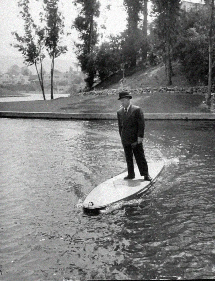 A man in a suit and hat stands on a small motorized surfboard, holding a handle, while gliding across a calm body of water near a shoreline with trees and houses in the background.