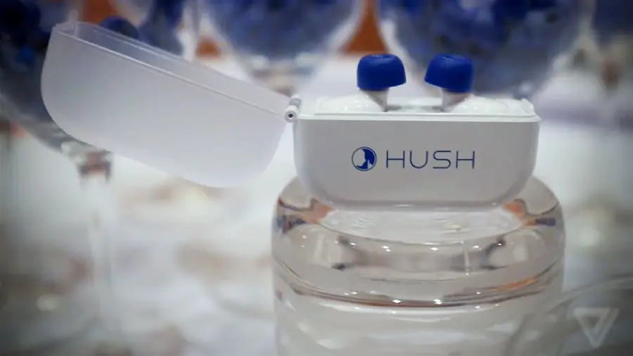 A white Hush earplugs case sits open, displaying blue-tipped wireless earplugs inside. The case is resting on a glass surface with blurred glasses in the background.