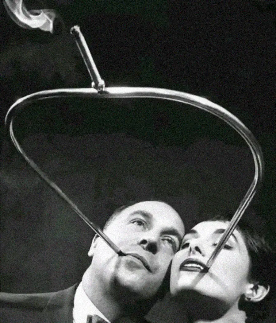 A man and a woman smile together as their cigarettes produce smoke that forms the shape of a heart around their faces against a dark background.