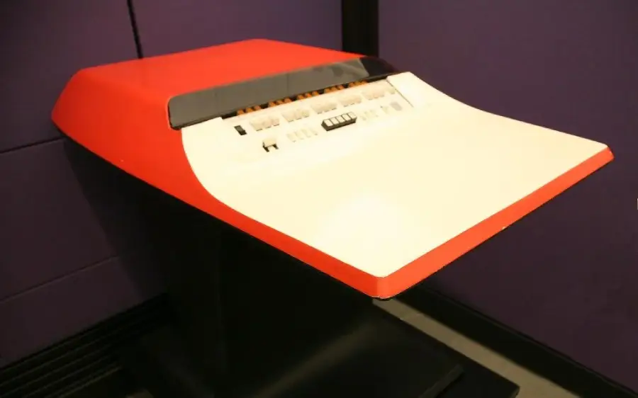 A vintage computer terminal with a red and white exterior, featuring a flat keyboard with various buttons and switches, set against a purple wall.