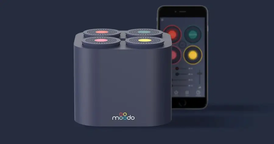 A dark gray Moodo smart aroma diffuser with four circular scent capsules on top is shown next to a smartphone displaying the Moodo control app.
