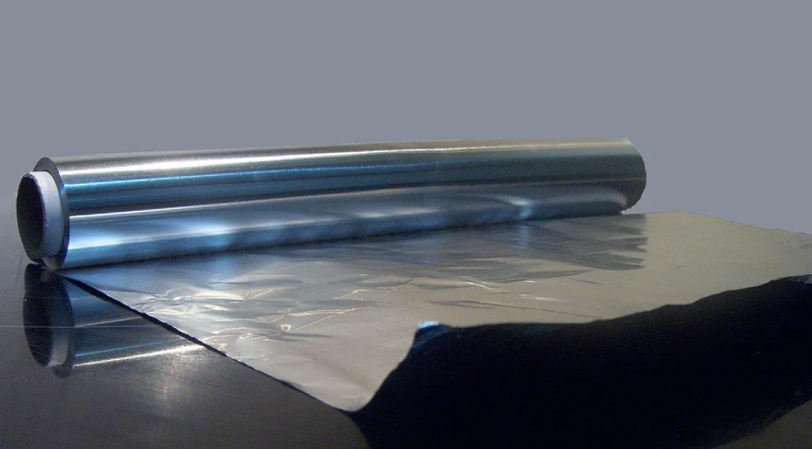 A roll of aluminum foil sits partially unrolled on a reflective surface, with a sheet of the shiny metal extending out from the roll. The background is plain gray.