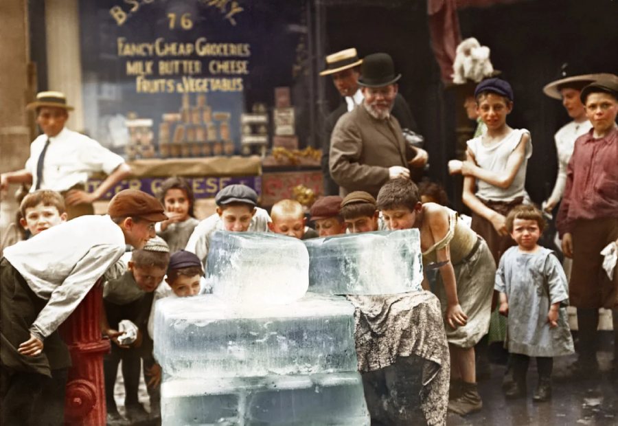 A group of children and a few adults gather around large blocks of ice on a city sidewalk in front of a grocery store, eagerly touching and cooling off in summer, early 20th century.