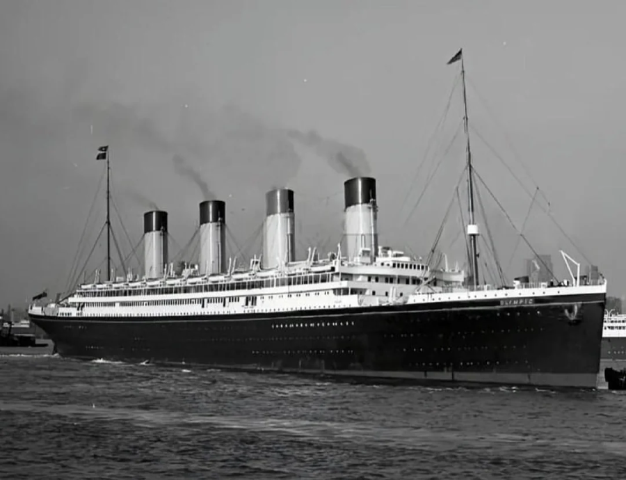 A large early 20th-century ocean liner with four smokestacks sails on calm water, emitting smoke. The ship is painted in dark and white colors and is seen from the side in a black-and-white photograph.